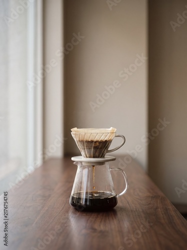 Manual Pour Over Coffee Brewing Process with Glass Dripper and Carafe on Wooden Table. Minimalist Morning Coffee Ritual in Soft Natural Window Light. Specialty Coffee Shop Atmosphere and Home Barista