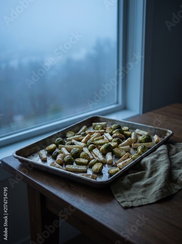 Roasted Brussels Sprouts and Parsnips on a Baking Sheet by a Window with Moody Blue Foggy Winter Landscape Background, Healthy Homemade Vegetable Side Dish Concept for Cozy Home Cooking and Nutrition