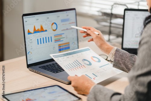 Businesswoman holding printed report and pointing with pen at laptop screen dashboard
