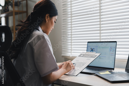 Female Asian doctor reviewing patient medical history clipboard document at office desk with laptop