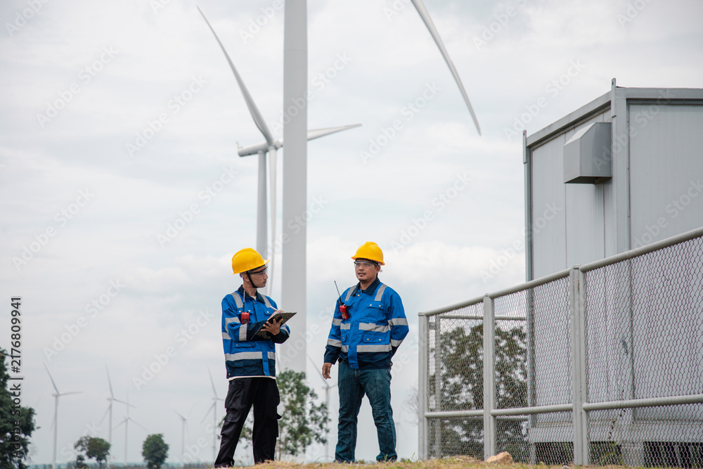 Fototapeta premium Two engineers in hard hats and safety gear reviewing documents on a clipboard at a wind turbine farm.