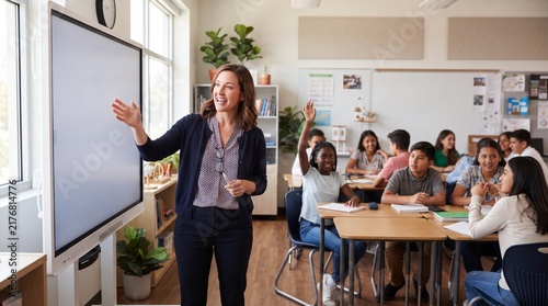 Female teacher conducting classroom lesson pointing at smartboard while diverse students listen