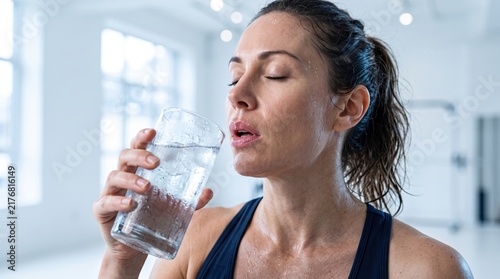 A tired woman taking a deep breath and drinking cold water after an intense workout session in a bright gym
