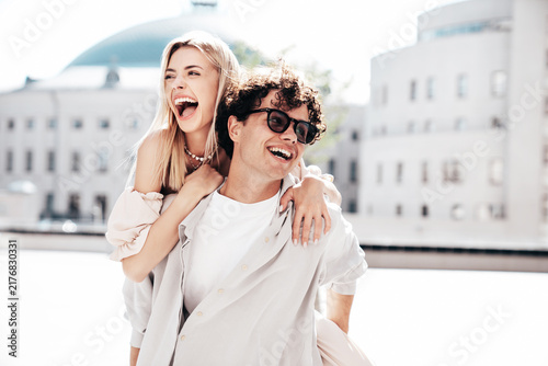 Young smiling beautiful woman and her handsome boyfriend in casual summer clothes. Happy cheerful family. Couple posing in street at sunny day. Female sits at his back. piggyback riding