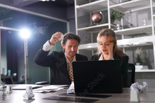 Konstfotografi Businessman lifts a crumpled ball ready to throw while the woman holds her temple, frustration and fatigue sharing the lamplit desk in a bright modern coworking office
