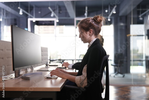 Bild auf Leinwand Young businesswoman with an auburn bun works at a widescreen desktop computer beside tall glass meeting rooms flooded with daylight in the open plan office in a bright modern coworking