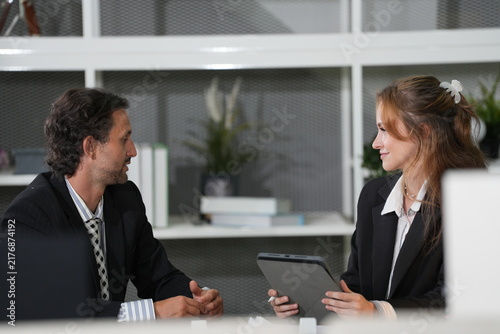 Foto Ginger-haired businesswoman lowers her gaze to the tablet while her long-haired colleague pauses, shelving with plants and books lining the wall behind in a bright modern coworking office