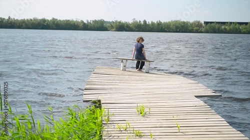 Girl sitting on a pier. Rear view Beautiful brunette girl with long hair sits on a wooden pier outdoors and looks into the distance while relaxing in nature.