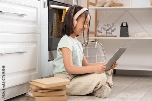 Little Asian girl with headphones and laptop studying online in kitchen