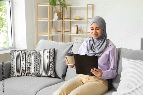 Muslim businesswoman with clipboard and coffee cup sitting on sofa at home