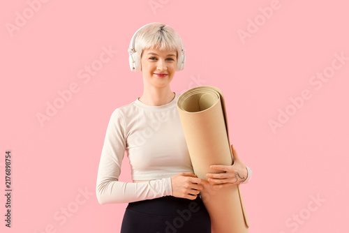 Sporty young woman in headphones with fitness mat on pink background