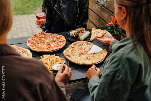 Group of friends enjoying pizza, fries, wraps and a drink together around an outdoor restaurant table in a casual social dining scene