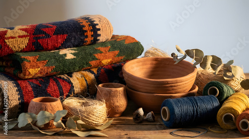 Handwoven artisan rugs stacked neatly beside natural clay pottery and colorful spools of thick thread with dried botanical leaves in warm golden hour lighting on a rustic wooden table surface.