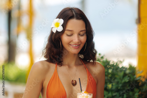 Young Woman with Plumeria Flower and Orange Swimsuit Drinking Cocktail