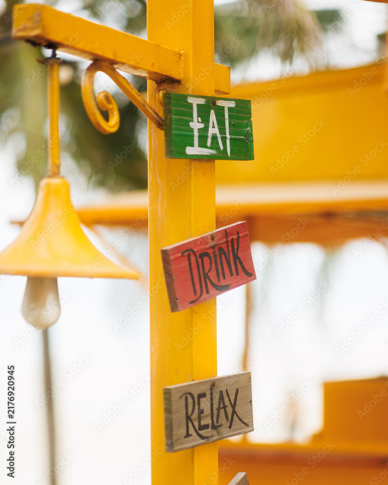 Obraz premium Wooden Eat Drink Relax Signs on Yellow Post with Bell Close-Up