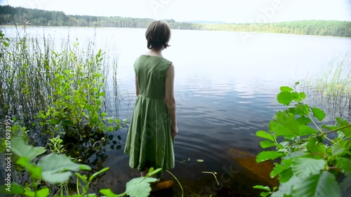 A young girl stands by a calm lake, enjoying nature. She wears a green dress that sways gently. The sun shines, creating a peaceful and joyful atmosphere.