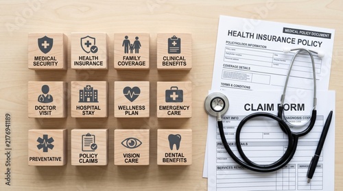 Wooden blocks display medical and health insurance terms alongside a health insurance claim form, pen, and stethoscope on a light wooden surface.