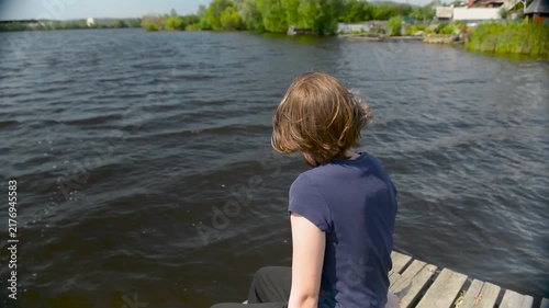 Girl sitting on a pier. Rear view Beautiful brunette girl with long hair sits on a wooden pier outdoors and looks into the distance while relaxing in nature.
