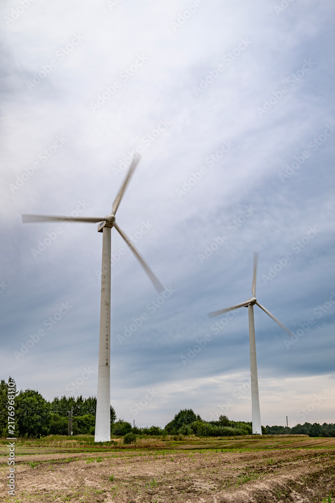 Fototapeta premium Low angle shot of two white wind turbines with spinning motion-blurred blades in an agricultural field under an overcast cloudy sky in Latvia.