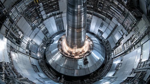 Inside a nuclear fusion reactor, a large central column emits light while surrounded by complex machinery and metallic structures