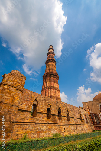 Tall red sandstone minaret tower rising above ancient stone wall ruin under blue sky with cloud at Qutb Minar, UNESCO World Heritage Site, Delhi, India.