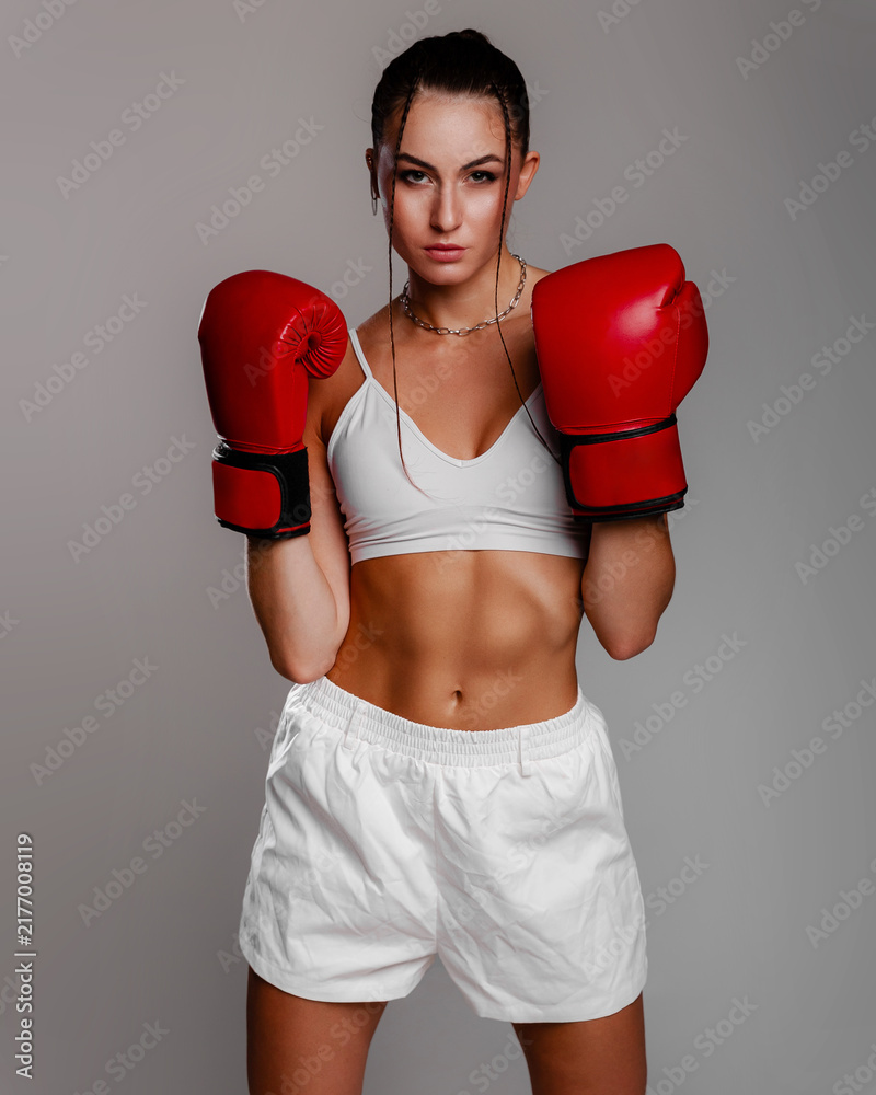 Obraz premium Athletic young woman in white sportswear and red boxing gloves poses confidently in a studio, creating a strong fitness concept for sports advertising, training campaigns and wellness media.