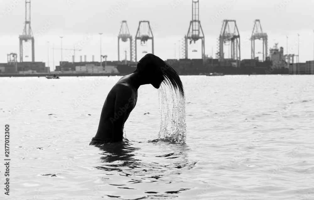 Obraz premium Silhouette of a young man flipping long hair back in sea water, port cranes background