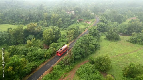 Aerial view of public red buses driving on rural asphalt road through lush green monsoon landscape.