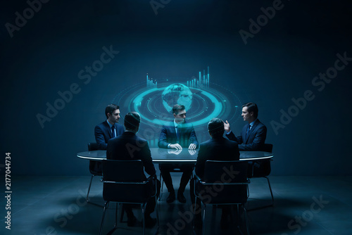 A group of business people sitting at a round table in a dark room, looking at a glowing blue holographic globe and data charts in the center