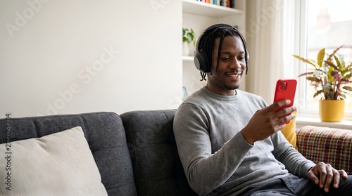 Happy African man with headphones using red phone on sofa at home.