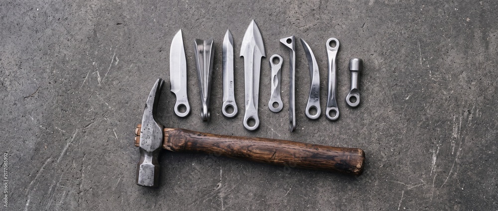 Collection of handmade metal forging tools including hammer and various metal shaped objects on textured concrete surface