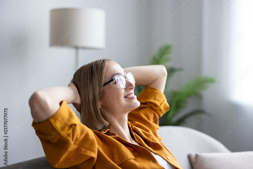 Obraz premium Young woman with eyeglasses relaxing on a sofa with hands behind her head, enjoying a moment of comfort and quiet happiness after achieving success at her remote work
