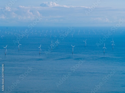 Offshore wind turbines standing across the sea off the North Wales coast with rows of wind generators stretching into the distance beneath visible clouds and blue sky over the water