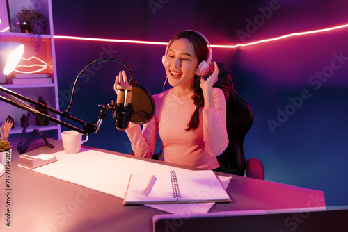 Obraz na płótnie Host channel of young beautiful Asian broadcaster listening the music on social media with listeners, wearing pastel headphones, using microphone for singing at neon light indoor studio
