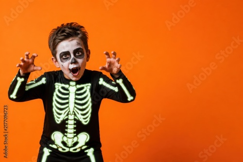 Scared child in skeleton costume with face paint on Halloween orange background with copy space