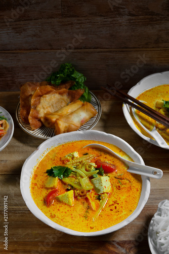 Creamy spicy Asian curry soup with tofu and vegetables, served with crispy spring rolls, pancakes, salad and chopsticks on a rustic wooden table.