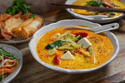 Creamy spicy Asian curry soup with tofu and vegetables, served with crispy spring rolls, pancakes, salad and chopsticks on a rustic wooden table.