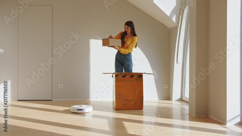 Woman in yellow shirt and jeans unboxing cardboard package with new robot vacuum cleaner on wooden floor