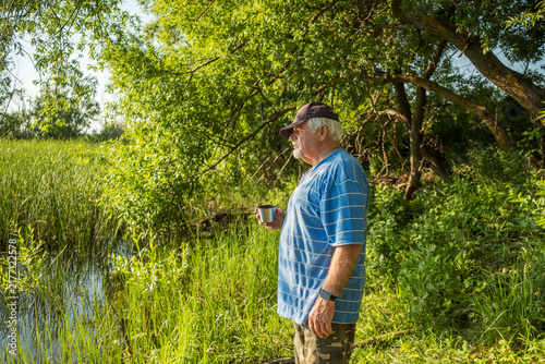Pensive aged male holding metal cup near river bank among green trees