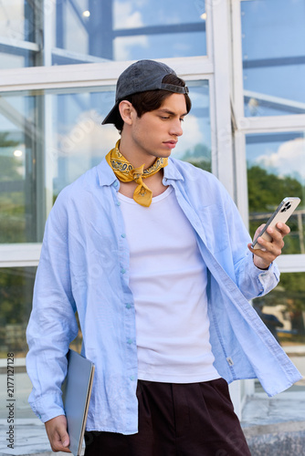 Bild auf Leinwand A young man checks his phone while enjoying the outdoor campus vibe in a blue sh
