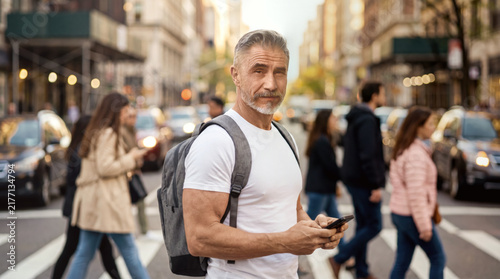 Gray haired man with backpack using smartphone on city crosswalk, urban traffic and pedestrians in background, focused modern lifestyle, mobile communication, outdoor portrait.