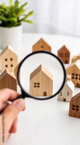 Hand Holding Magnifying Glass Over Small Wooden Toy House Models On White Surface, Real Estate Search Concept