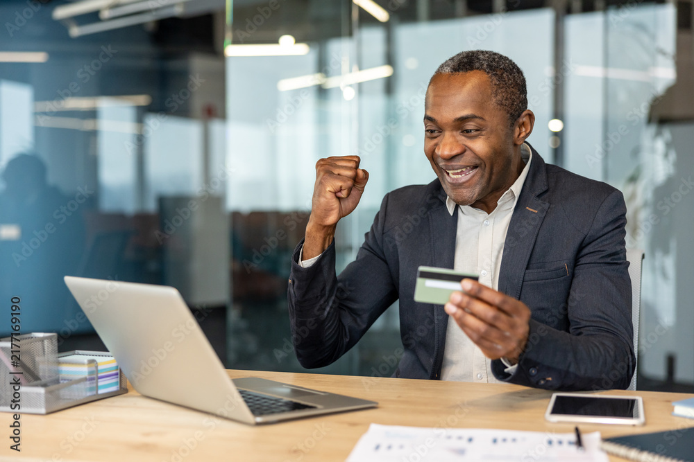 Obraz premium Mature businessman receiving good news on a laptop, holding a credit card and expressing joy and excitement at his desk in a modern office, symbolizing achievement and successful online transaction