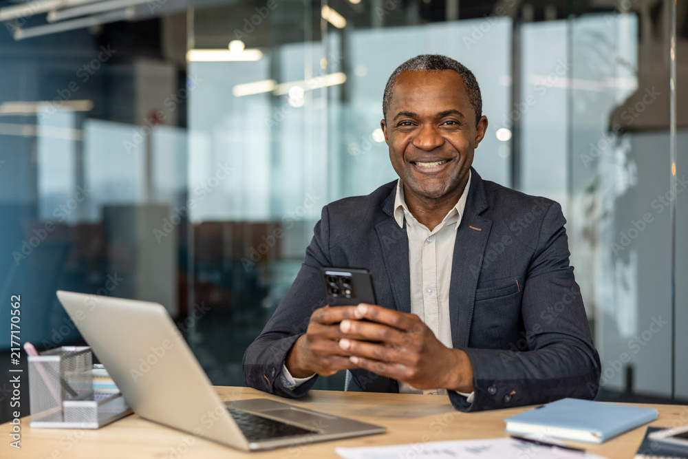 Obraz premium Mature professional manager engaging with technology in a modern corporate office, confidently communicating and enjoying a successful workday while using a mobile device at his desk