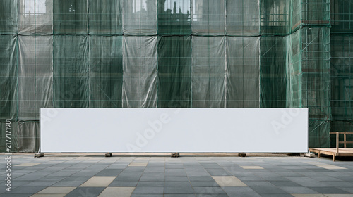 A long blank white billboard or fence mockup set against a building facade covered with green protective scaffolding mesh and a tiled pavement.