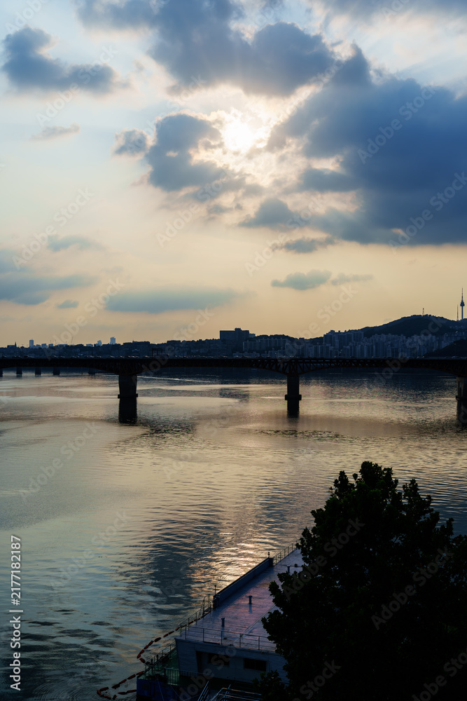 Obraz premium Sunset over river bridge and city skyline with cloudy sky