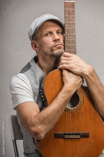 Close portrait of middle-aged man holding acoustic guitar upright, cheek resting on fretboard, calm thoughtful expression with neutral studio background and warm tones, showing adult hobby and quiet