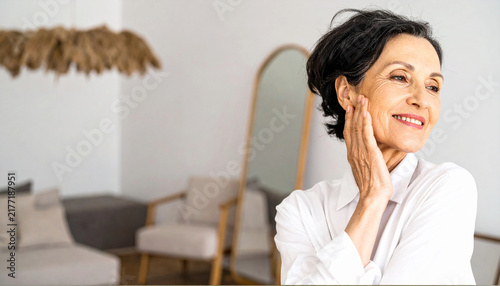 Confident middle aged woman touching her face with gentle smile and natural makeup indoors