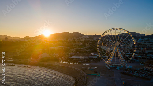 aerial drone shot of a Sunrise over Marseille’s waterfront with Ferris wheel, calm shoreline, rocky jetty, and golden mountains creating a serene coastal cityscape