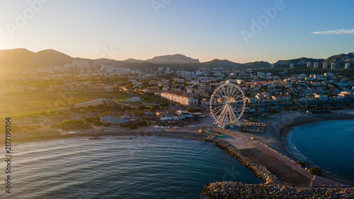 aerial drone shot of a Sunrise over Marseille’s waterfront with Ferris wheel, calm shoreline, rocky jetty, and golden mountains creating a serene coastal cityscape
